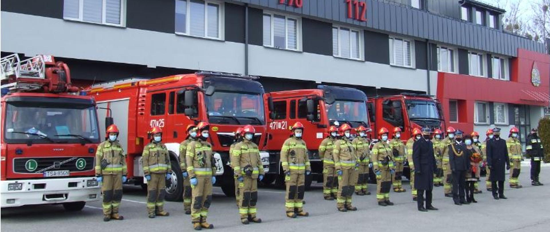 Zdjęcie przedstawia stojących w dwuszeregu strażaków na tle budynku strażnicy i samochodów strażackich. Strażacy są ubrani w ubrania specjalne, na głowach mają hełmy. Przed dwuszeregiem stoi trzech strażaków w mundurach galowych.