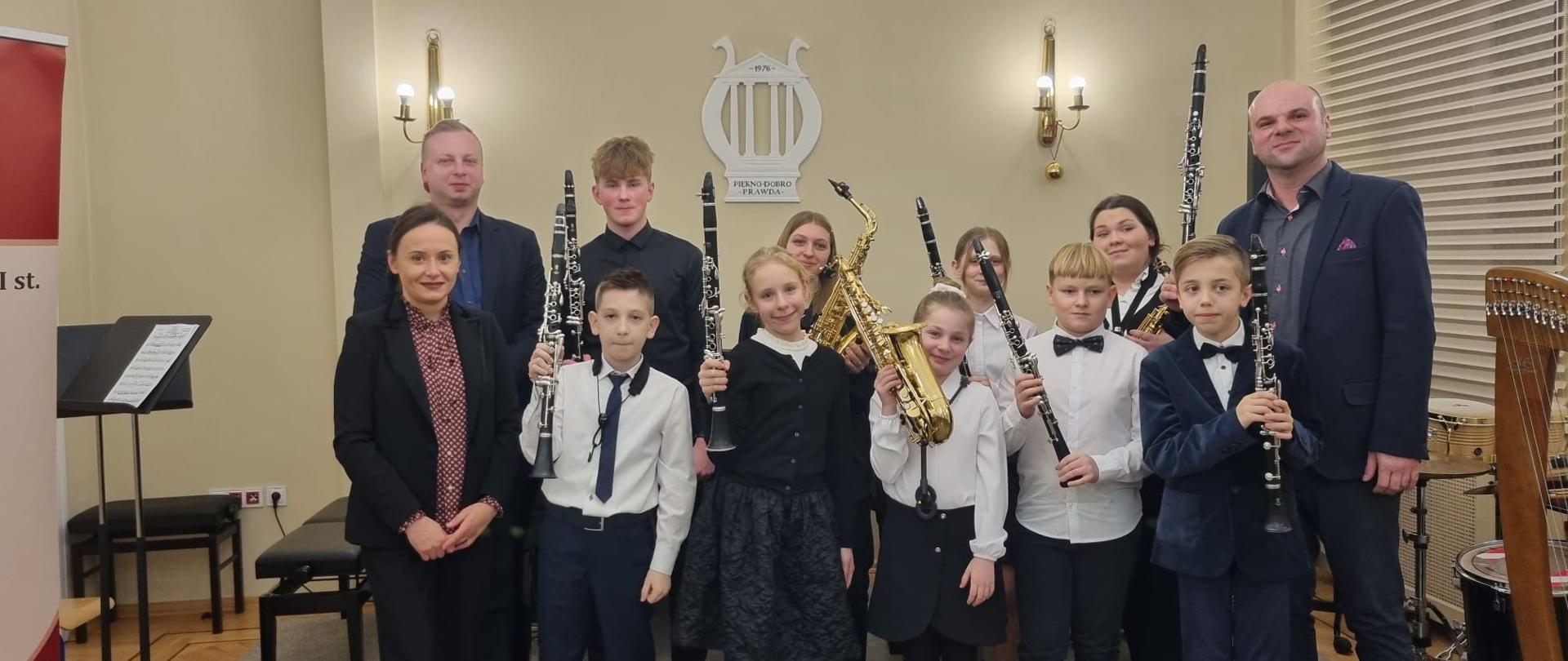 Uczniowie i nauczyciele. Zdjęcie wykonane w auli szkoły. Uczniowie trzymają w dłoniach saksofony i klarnety.