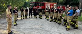 Zbiórka ćwiczących strażaków po ćwiczeniach. W tle stoi pojazd pożarniczy z przyczepą.