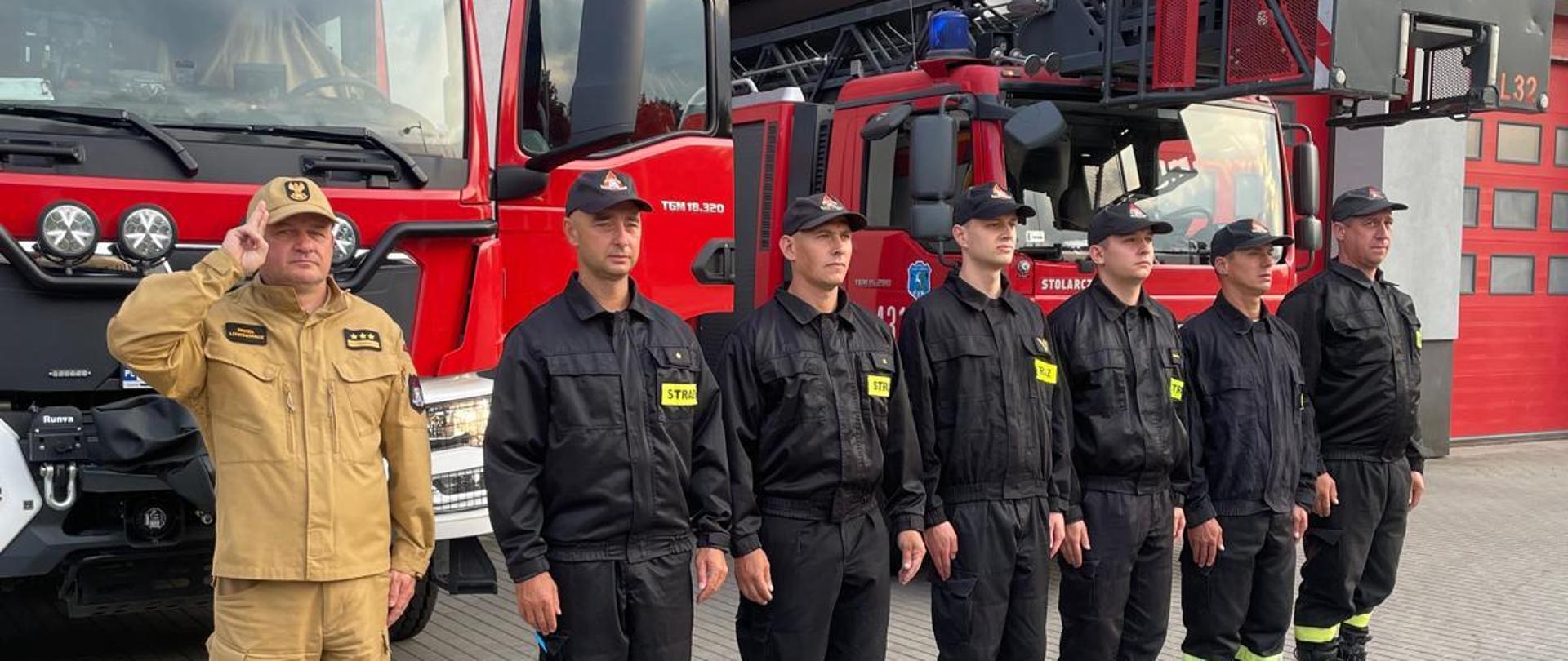 Zdjęcie przedstawia 7 strażaków oddających hołd zmarłym strażakom, w tle Komenda oraz pojazdy pożarnicze