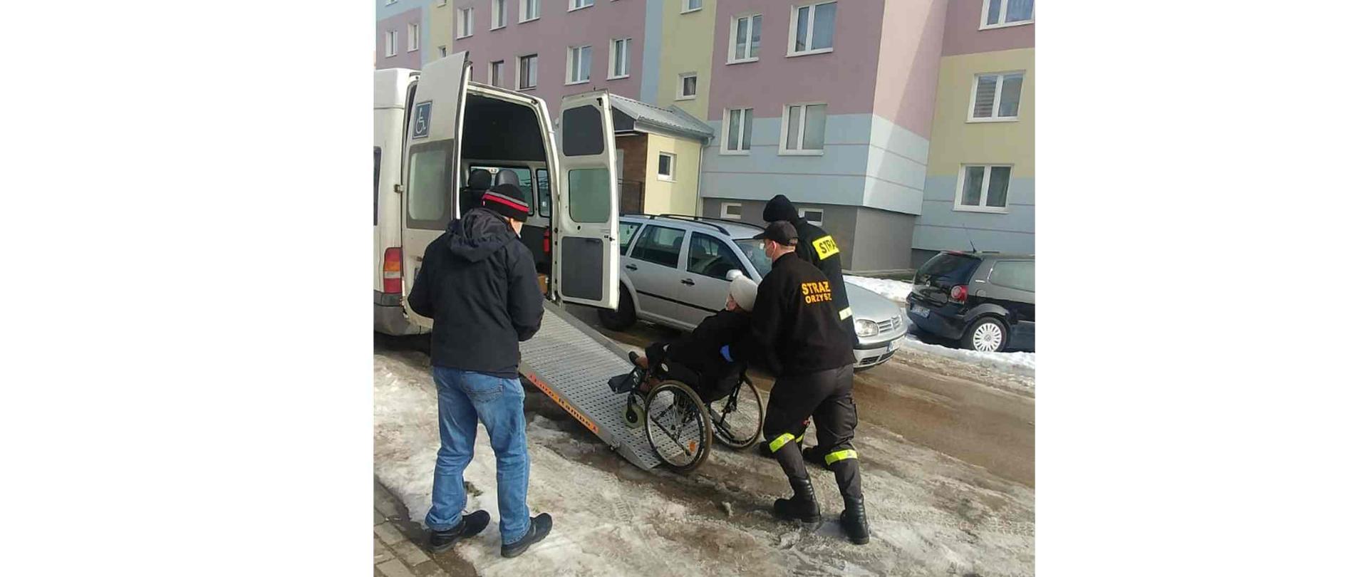 Dwóch strażaków wnoszących kobietę na wózku inwalidzkim do samochodu.