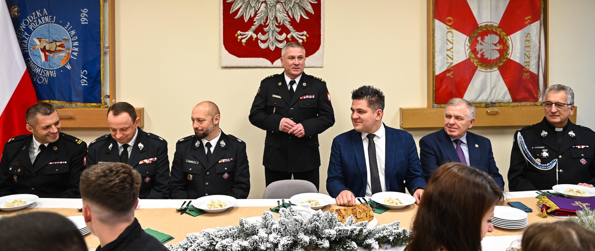 na zdjęciu widoczny Komendant Miejski w trakcie przemowy, obok zaproszeni goście,
w tle godło Rzeczypospolitej Polskiej oraz wiszące na ścianie poczty sztandarowe
