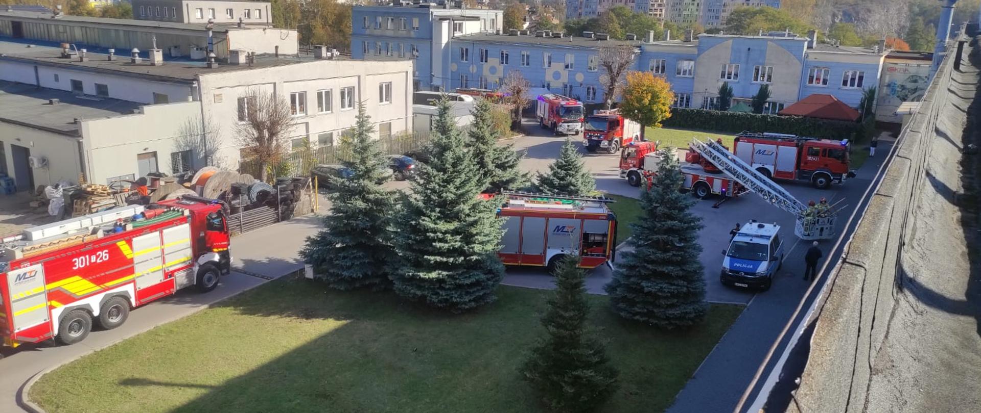 Zdjęcie przedstawia teren hali produkcyjnej przy ulicy Mielczarskiego (widok z dachu) gdzie stoi 7 wozów strażackich i policyjny radiowóz.