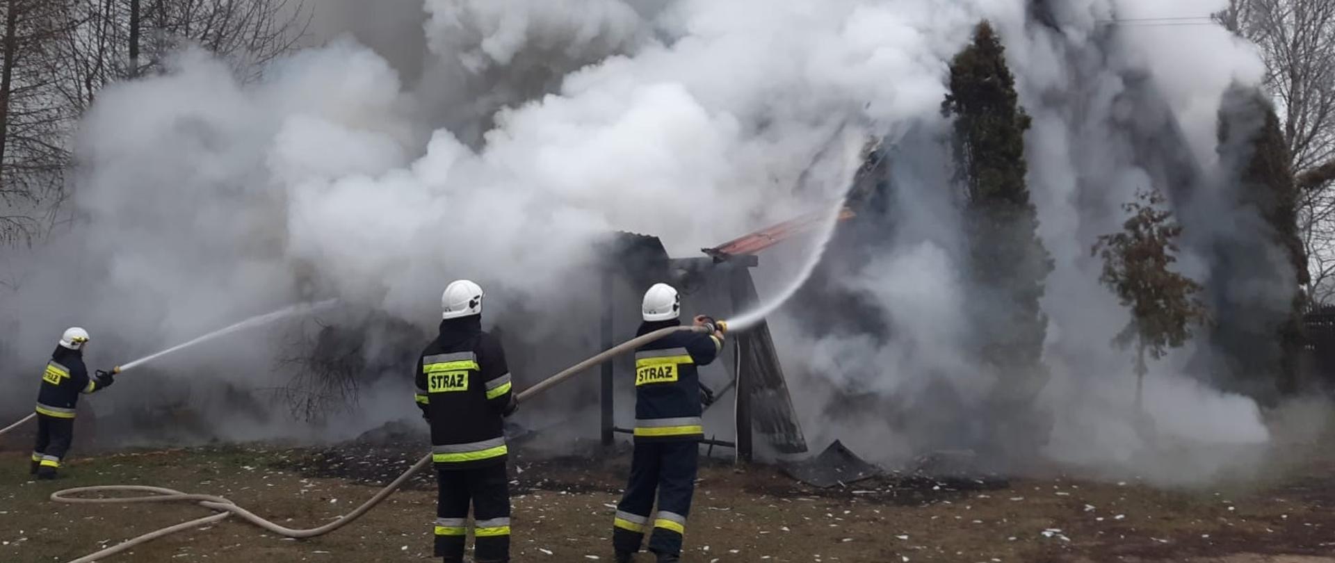 Zdjęcie obrazuje 3 strażaków lejących wodę z węży na palący się budynek, który jest objęty kłębami dymu. 