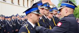 Widok z boku. Zastępca Komendanta Wojewódzkiego w trakcie dekoracji odznaczeniami zasłużonych funkcjonariuszy. W tle pododdział OSP. 