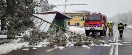 Widok na drogę asfaltową. Lewy pas zablokowany przez powalone drzewo. Za przewróconym drzewie przystanek autobusowy. Obok niego stoi samochód strażacki. Po prawej stronie widocznych trzech strażaków w ubraniach specjalnych i hełmach. 