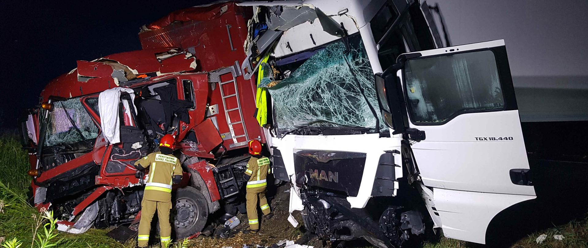 Zdjęcie zrobione w nocy na drodze krajowej Dk 9. Na zdjęciu widać dwa rozbite samochody ciężarowe. Obok jednego z nich stoi dwóch strażaków wykonujących swoje czynności. W oddali widać porozrzucane elementy samochodów. 