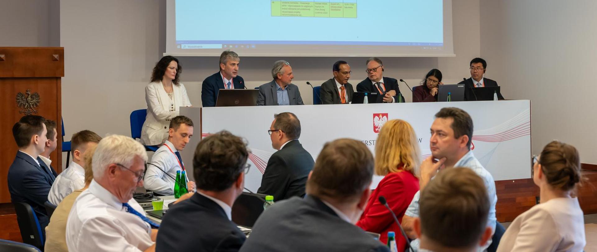 Wiceminister finansów Jarosław Neneman podczas spotkania misji Międzynarodowego Funduszu Walutowego, siedzi za stołem z innymi prelegentami, z przodu siedzą inni uczestnicy spotkania