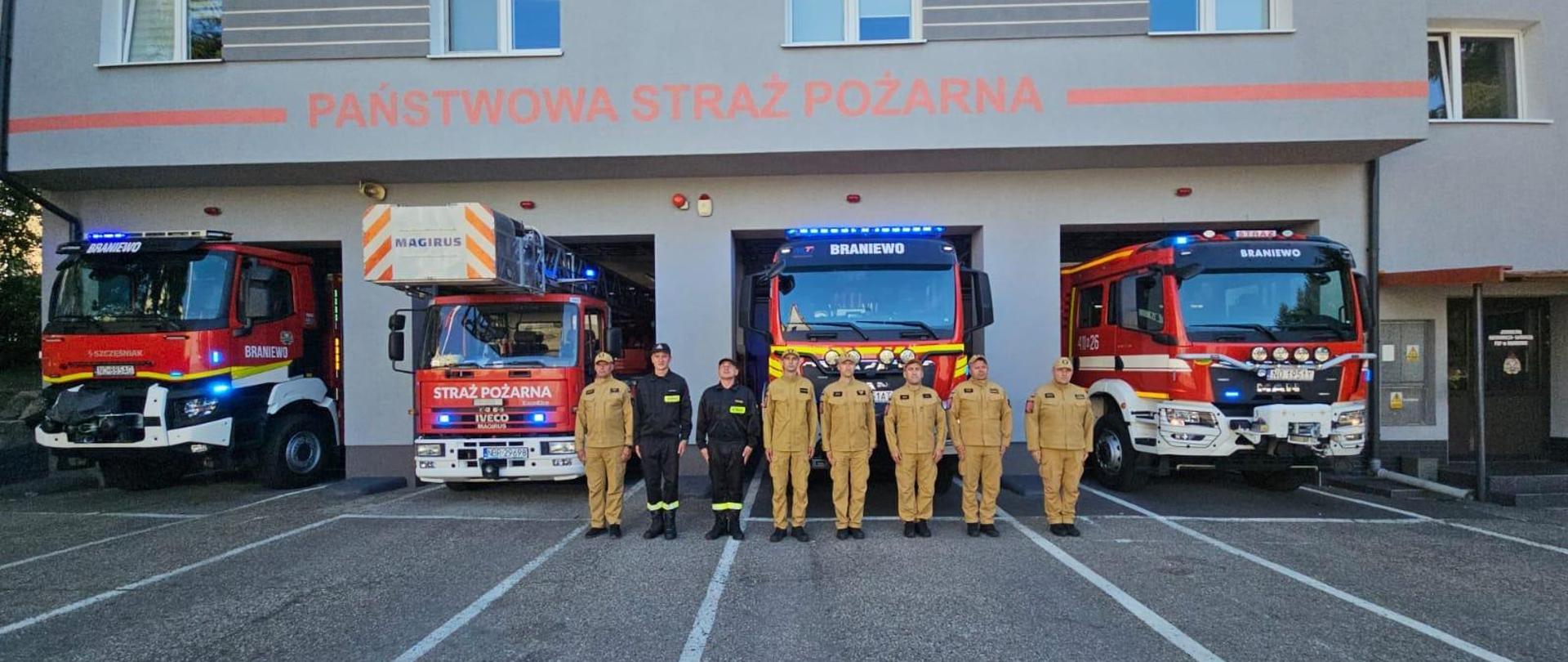 Strażacy w mundurach ustawieni w jednym szeregu na tle czterech samochodów strażackich i remizy jednostki ratowniczo gaśniczej.