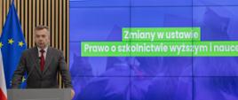 Minister Wieczorek stoi za mównicą i mówi do mikrofonu, za nim na wbudowanym w ścianę ekranie napis Zmiany w ustawie Prawo o szkolnictwie wyższym i nauce.