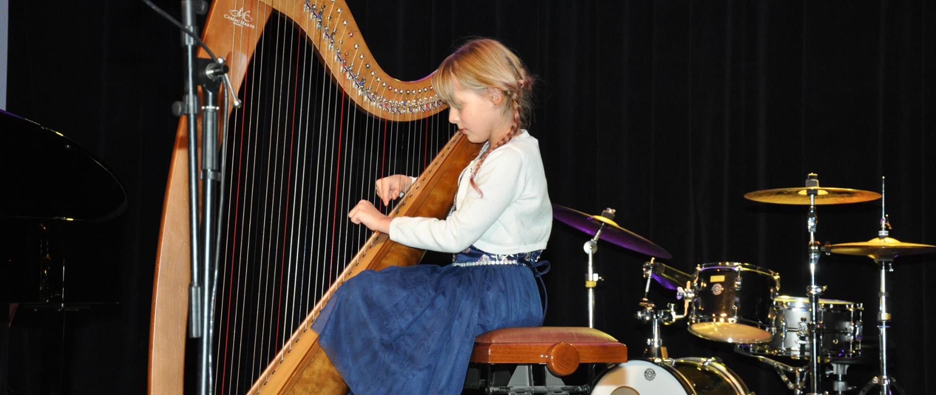 Dziewczynka grająca na harfie. W tle instrumenty perkusyjne.