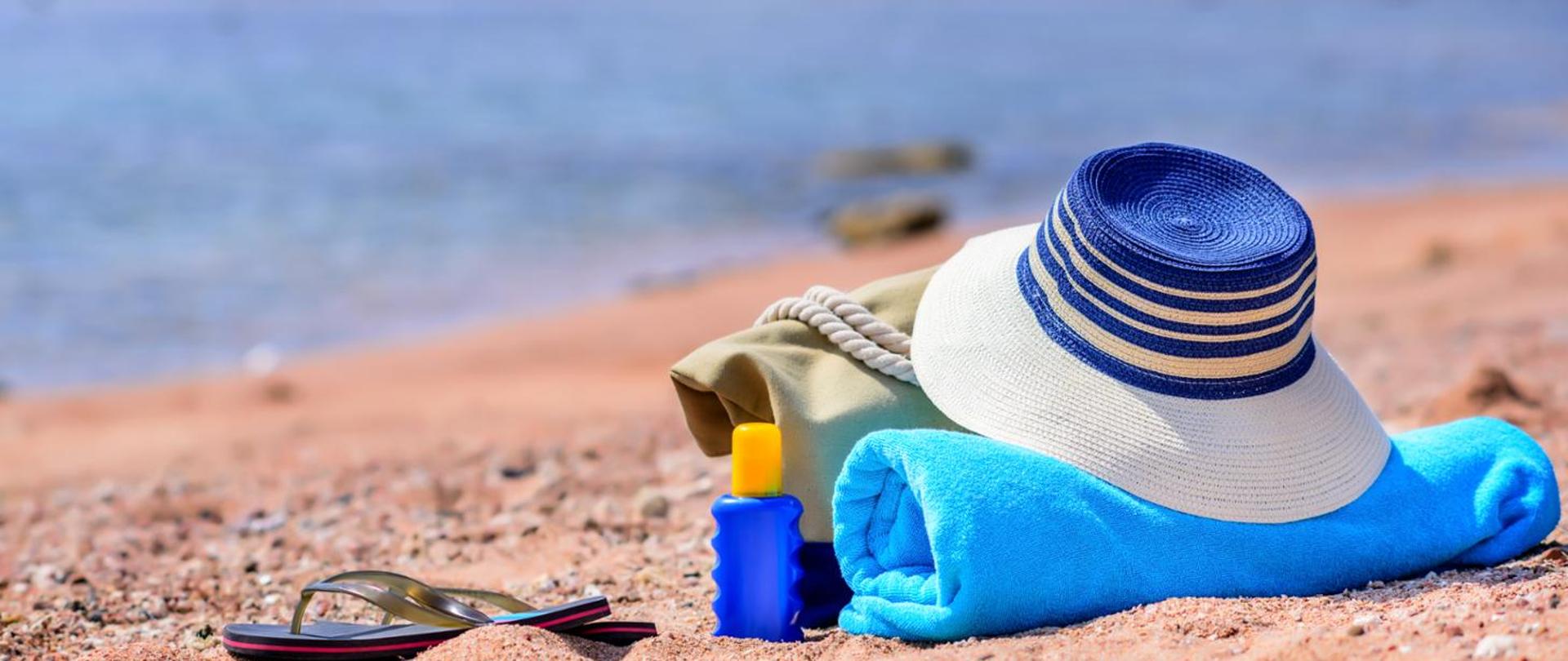 Na piaszczystej plaży leżą: niebieski ręcznik, biało-niebieski kapelusz, olejek do ciała oraz klapki kąpielowe i koc