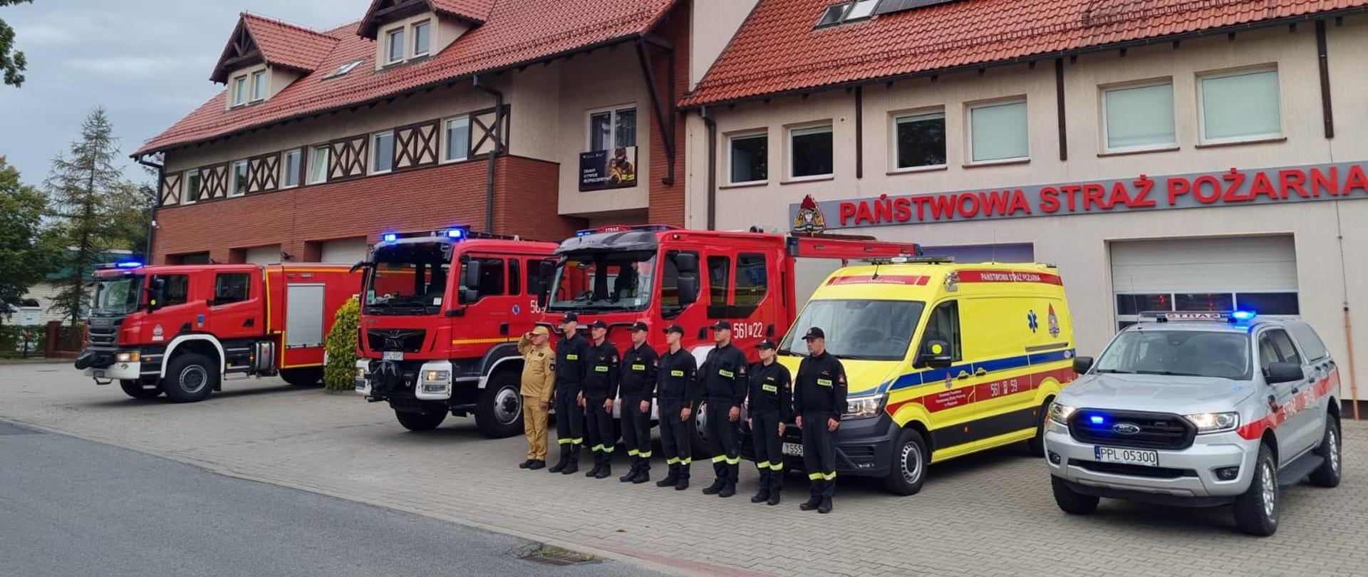 Strażacy stojący w szeregu oddają hołd poległym strażakom. W tle samochody pożarnicze oraz budynek strażnicy.