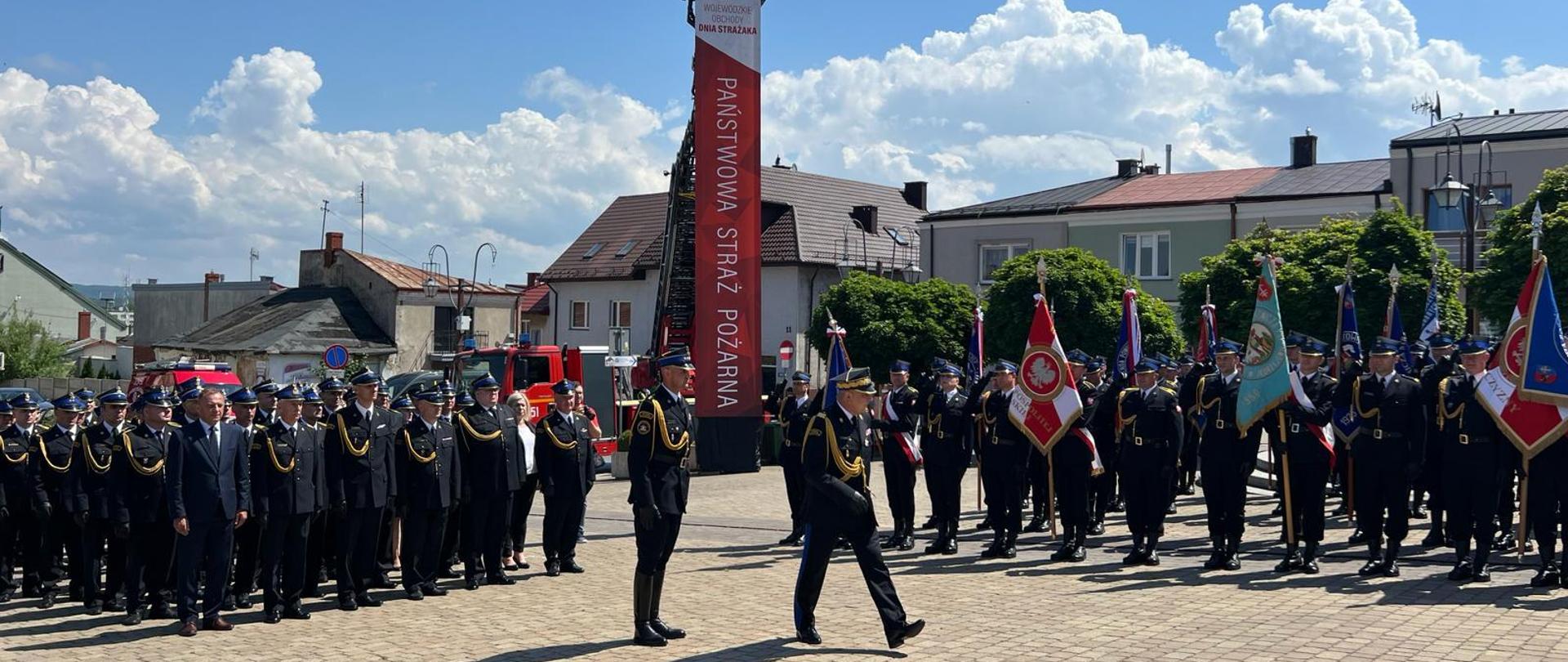 Zastępca komendanta głównego Państwowej Straży Pożarnej dokonuje przeglądu pocztów sztandarowych i pododdziałów podczas wojewódzkich obchodów dnia strażaka województwa świętokrzyskiego. Za pododdziałami stoi autodrabina strażacka z banerem Państwowej straży Pożarnej. 