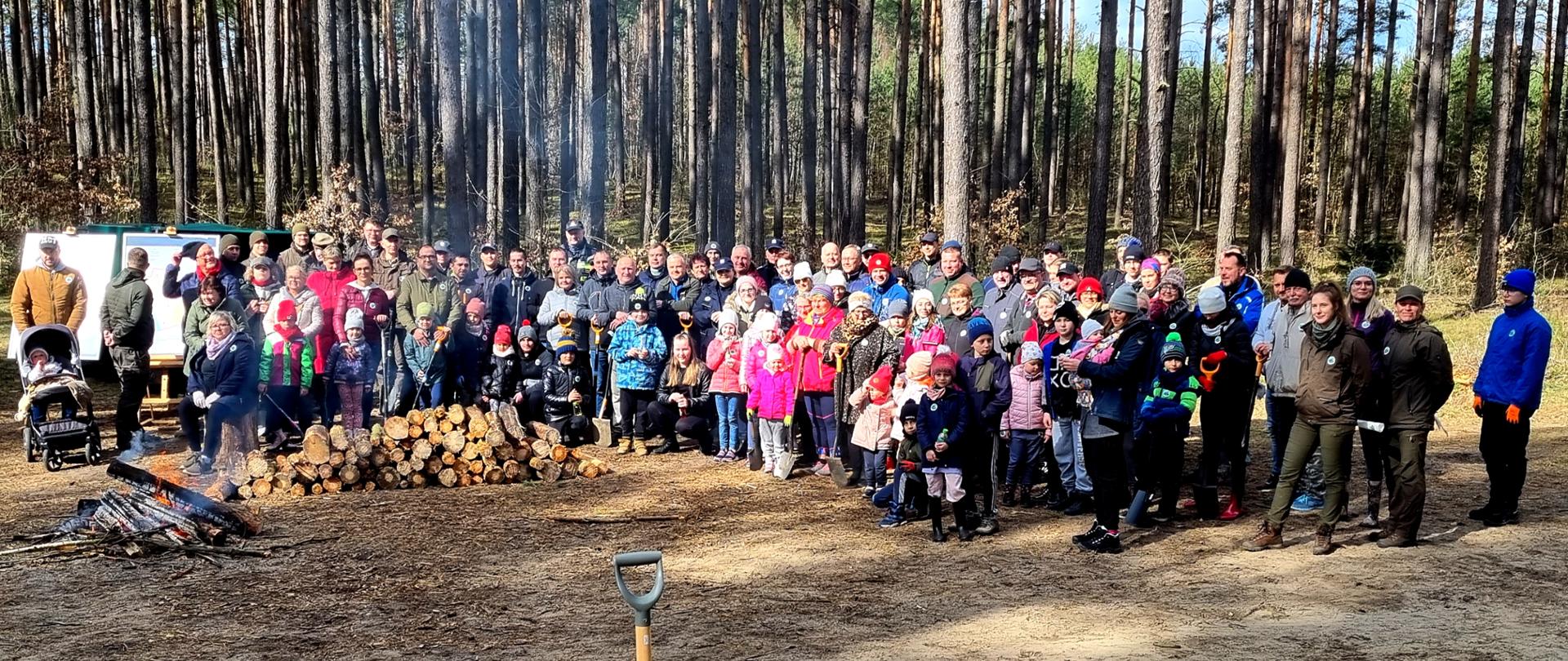 Zdjęcie przedstawia wszystkich uczestników akcji na tle lasu, widać dzieci i dorosłych, ognisko i drzewo do palenia.