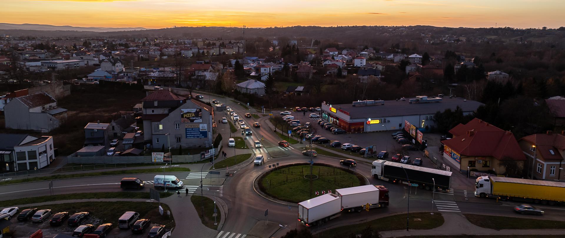 Widok z góry na rondo na DK73 w Jaśle. Widać auta stojące w korku na każdym wlocie na rondo. Są to auta osobowe i ciężarowe. POra popołudniowa, widać powoli zmierzch. 