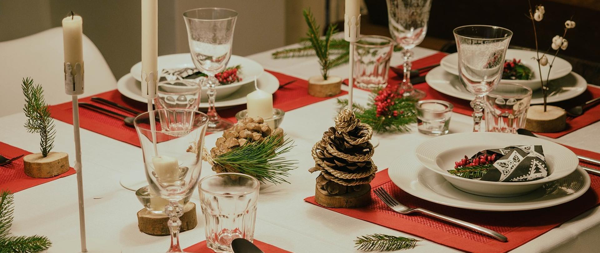 Stół wigilijny zaścielony białym obrusem, wraz z zastawą stołową w kolorze białym, na czerwonych podkładkach. Na stole znajdują się dekoracje w postaci szyszek i zielonych igieł świerka