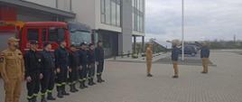 Zdjęcie przedstawia strażaków na placu przed Komendą. Po lewej stronie w rzędzie stoją strażacy , za nimi czerwony samochód, na środku dowódca melduje Zastępcy Komendanta Wojewódzkiego. Po prawej stronie stroi Komendant Powiatowy