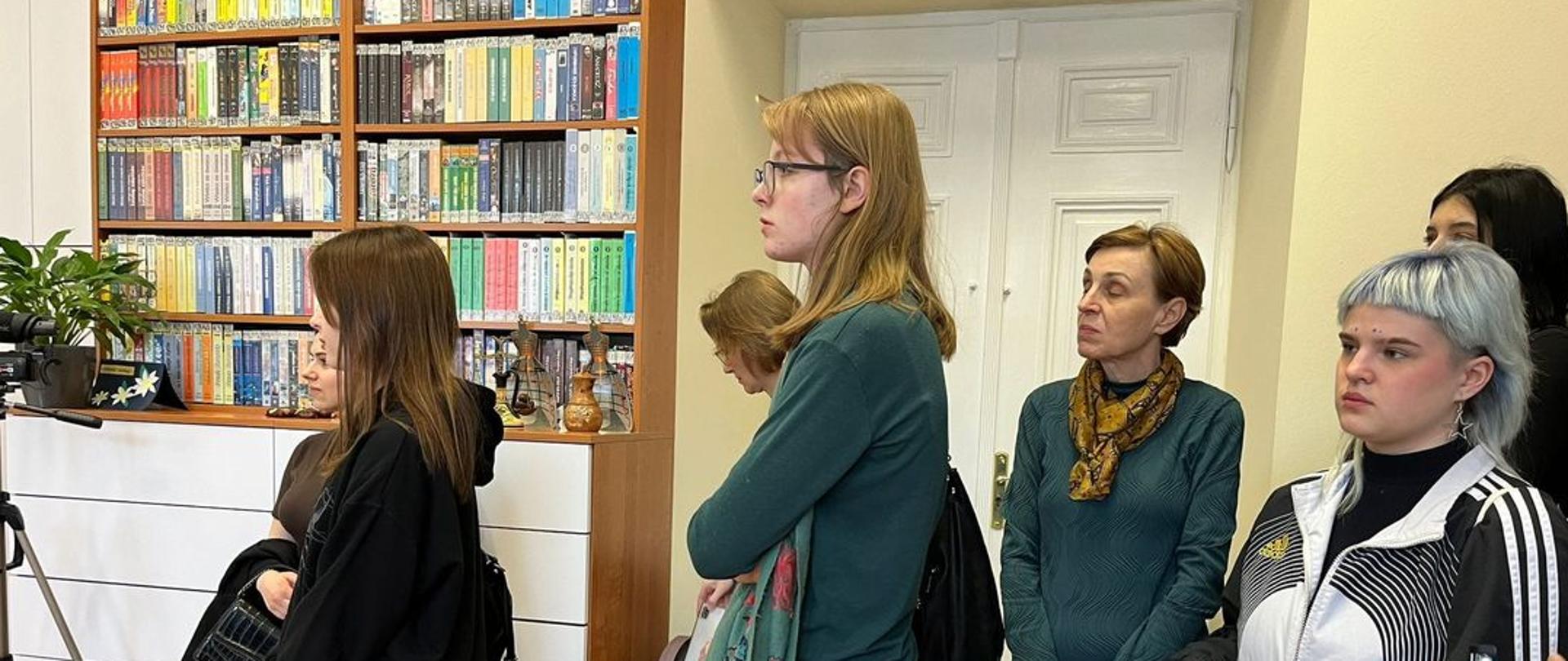Uczniowie i Jolanta Jarosińska słuchają w PWBP w Lublinie. Stoją na tle białej komody, regałów z książkami i białych, stylowych drzwi.