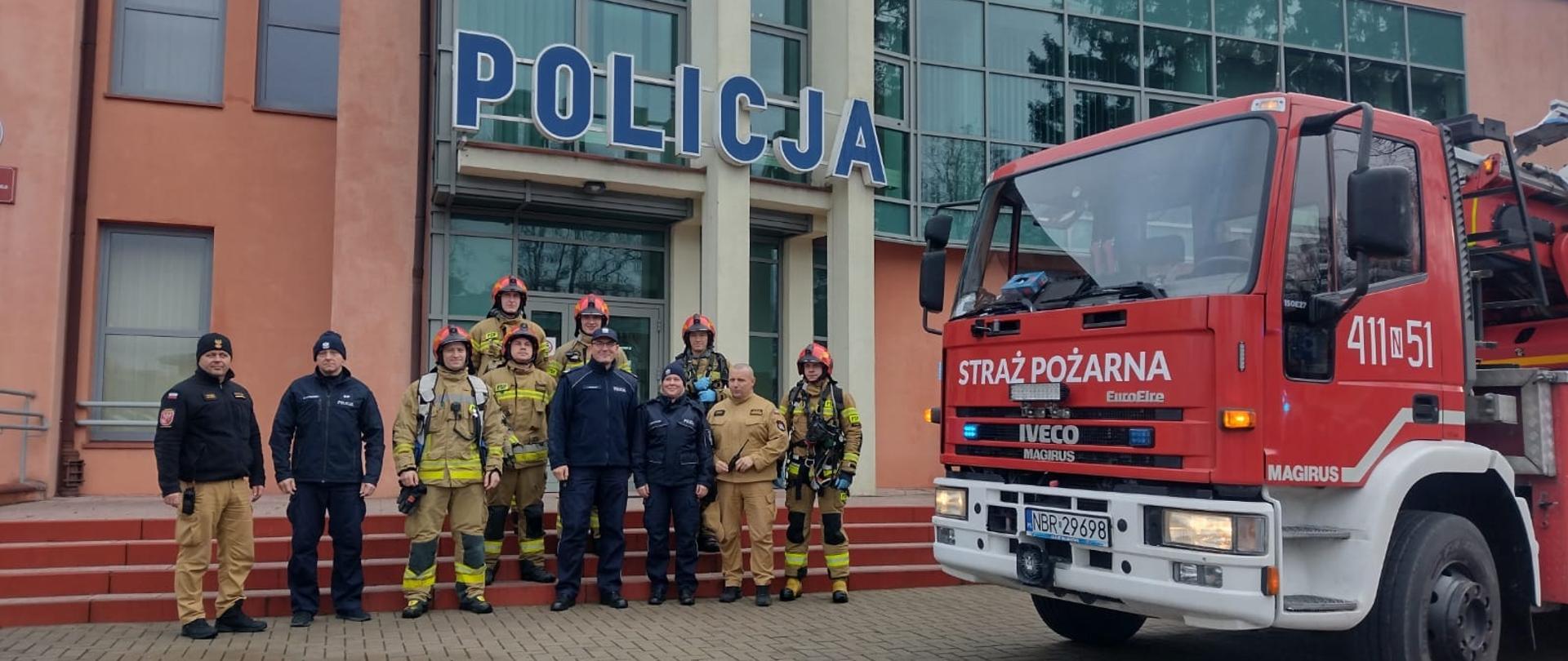 Przed wejściem do budynku komendy policji stoją strażacy i funkcjonariusze policji.