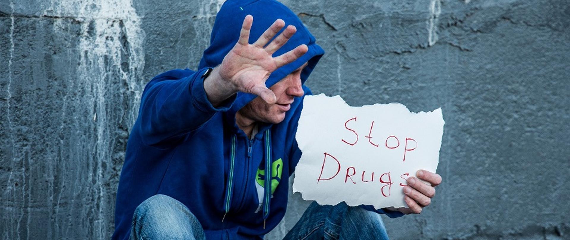 Siedzący na ziemi mężczyzna pod ścianą, w niebieskiej bluzie z kapturem na głowie. Prawą rękę ma wyciągniętą do przodu i dłonią pokazuje symboliczny znak "Stop", a w lewej ręce trzyma kartkę z napisem "Stop drugs"