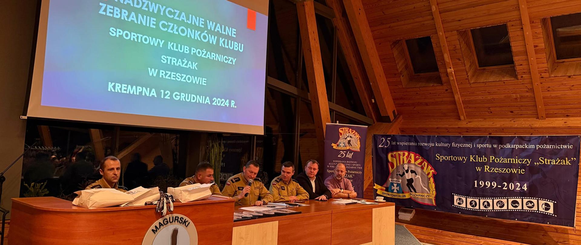 Na zdjęciu widać salę konferencyjną z drewnianym wykończeniem, w której odbywa się nadzwyczajne walne zebranie członków klubu „Sportowy Klub Pożarniczy Strażak” z Rzeszowa. Na środku sali stoi stół prezydialny, przy którym siedzi sześciu mężczyzn w mundurach strażackich, z dokumentami i mikrofonami przed sobą. Za stołem znajduje się duży ekran projekcyjny wyświetlający napis:
„NADZWYCZAJNE WALNE ZEBRANIE CZŁONKÓW KLUBU
SPORTOWY KLUB POŻARNICZY STRAŻAK W RZESZOWIE
KREMNPA 12 GRUDNIA 2024 R.”
Po prawej stronie ekranu stoi baner z logotypem klubu oraz informacją o 25-leciu działalności (1999-2024), podkreślający wsparcie dla rozwoju kultury fizycznej i sportu w podkarpackim pożarnictwie. W tle widać drewniane belki konstrukcyjne i oświetlenie sufitowe. Na przodzie stołu umieszczone jest logo „Magurski Park Narodowy”