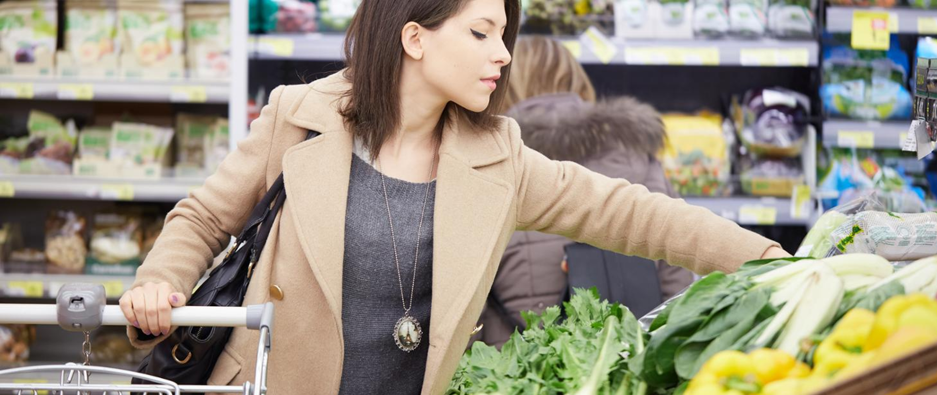 na pierwszym planie kobieta trzymająca rękę na poręczy wózka sklepowego, druga ręka sięga po warzywa. W tle półki sklepowe z produktami.