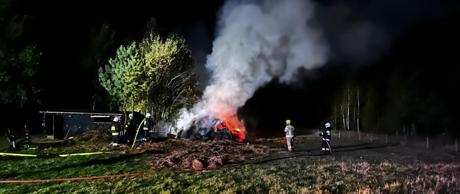 Zdjęcie przedstawia baloty siana w ogniu. Z dwóch stron płonącej pryzmy znajdują się strażacy: z lewej podają wodę z prawej kontrolują pożar.