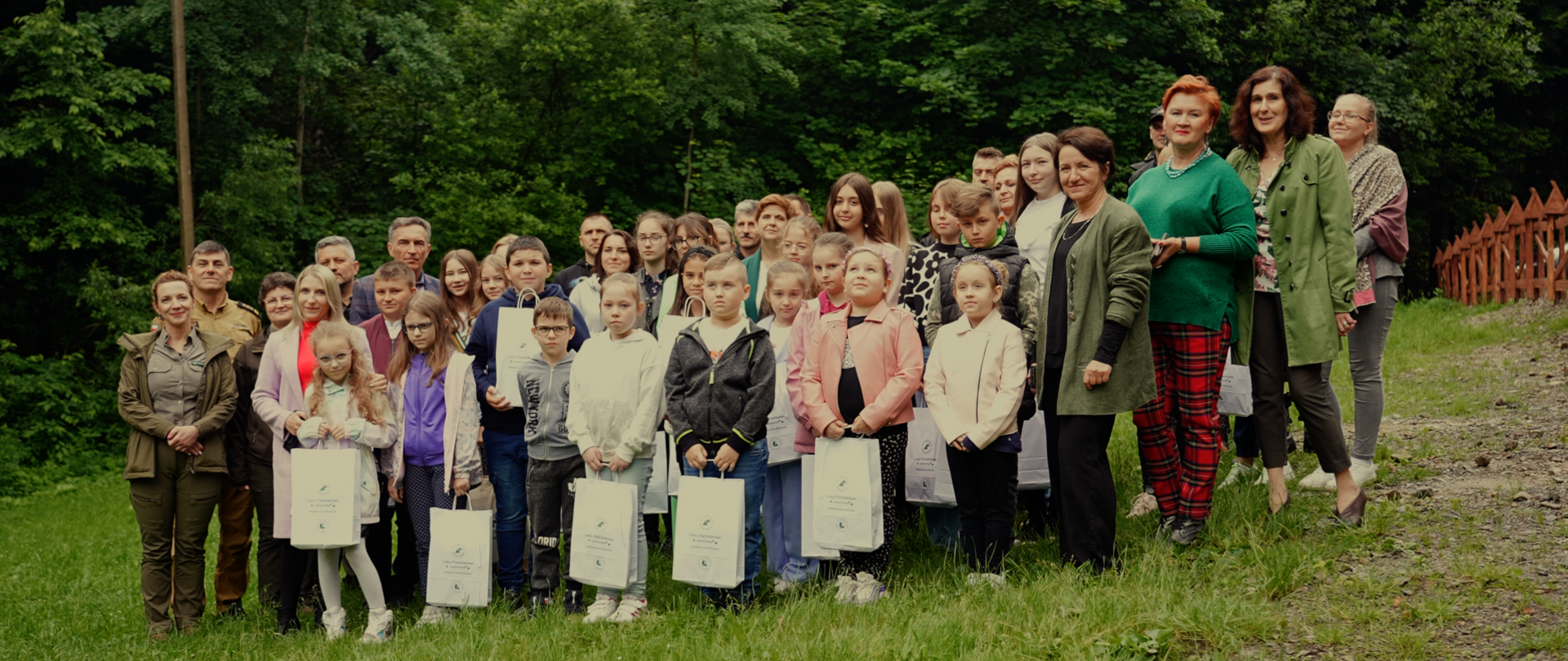 Kolorowa fotografia wykonana na zewnątrz w pochmurny dzień. Przedstawia laureatów konkursu plastycznego wraz z nauczycielami i opiekunami oraz osobami biorącymi udział w rozstrzygnięciu konkursu. Fotografia wykonana na tle lasu.