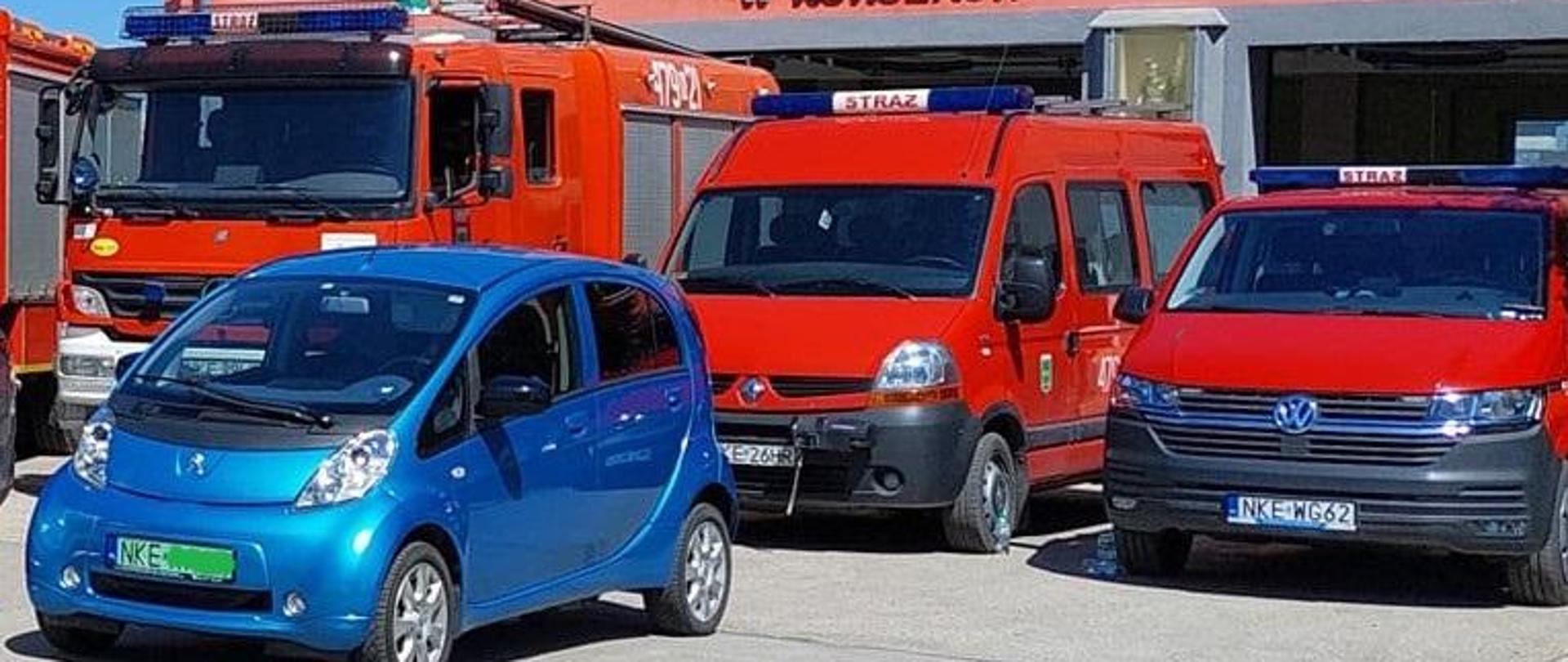 Na zdjęciu widać pojazd osobowy, niebieski i pojazdy straży pożarnej czerwone. W tle budynek remizy.
