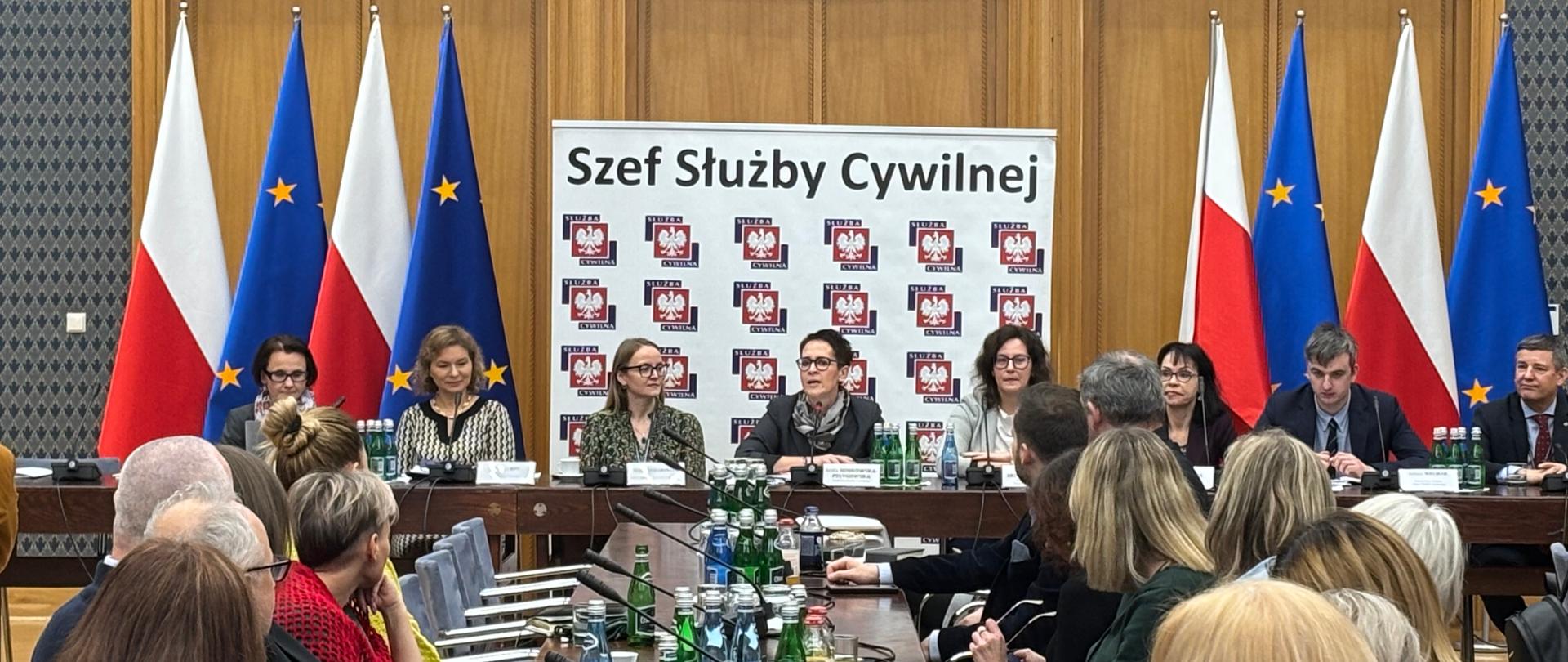 Za stołem konferencyjnym siedzi osiem osób ( 6 kobiet i 2 mężczyzn). W tle baner z logo szefa służby cywilne. Po lewej i prawej stronie od baneru flagi Polski i UE. Kobieta przy stole konferencyjnym przemawia do mikrofonu. Na pierwszym planie uczestnicy konferencji ( na zdjęciu widoczny jeden stół)