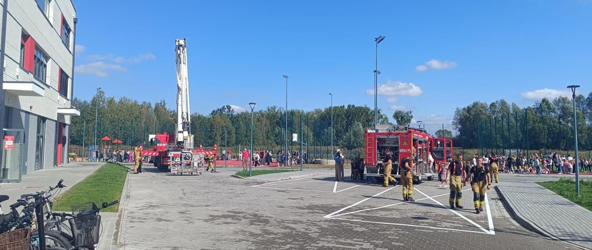 Samochody strażackie stoją na parkingu z kostki koło szkoły podnośnik jest rozstawiony. Pomiędzy samochodami chodzą strażacy. Na trawie za samochodami w grupach stoją dzieci i nauczyciele po próbnej ewakuacji. 