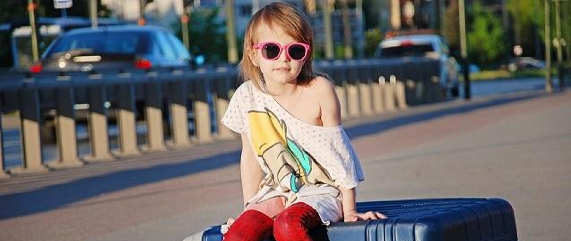 dziewczynka w okularach słonecznych siedząca na walizce na chodniku