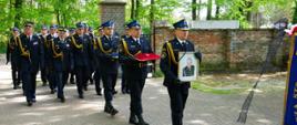 Kondukt pogrzebowy wchodzi na cmentarz , strażak prowadzący niesie w rękach portret strażaka zmarłego, kolejny niesie poduszkę czerwoną z medalem a kolejny czapkę rogatywkę