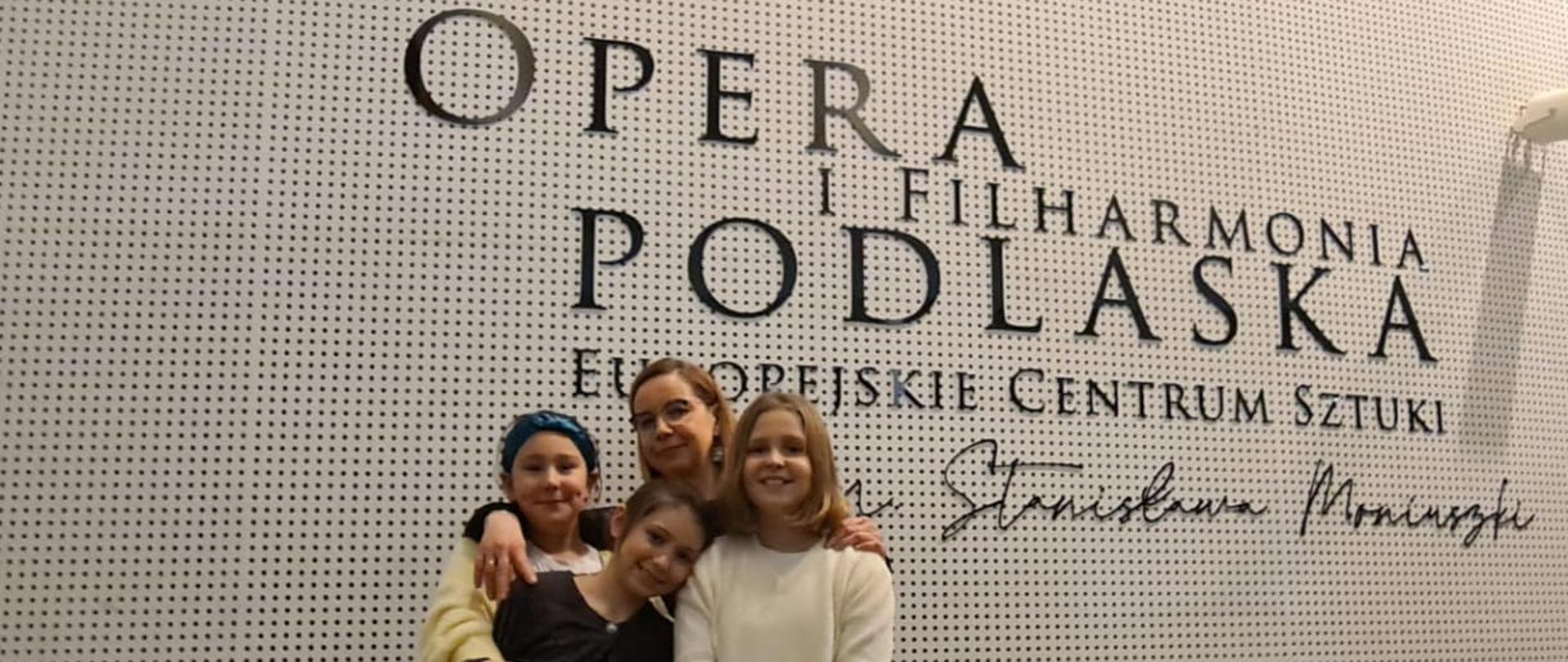 Zdjęcie przedstawia trzy uczennice i ich nauczycielkę, stojące na tle białej ściany z logo Opery i Filharmonii Podlaskiej - Europejskiego Centrum Sztuki im. S. Moniuszki.