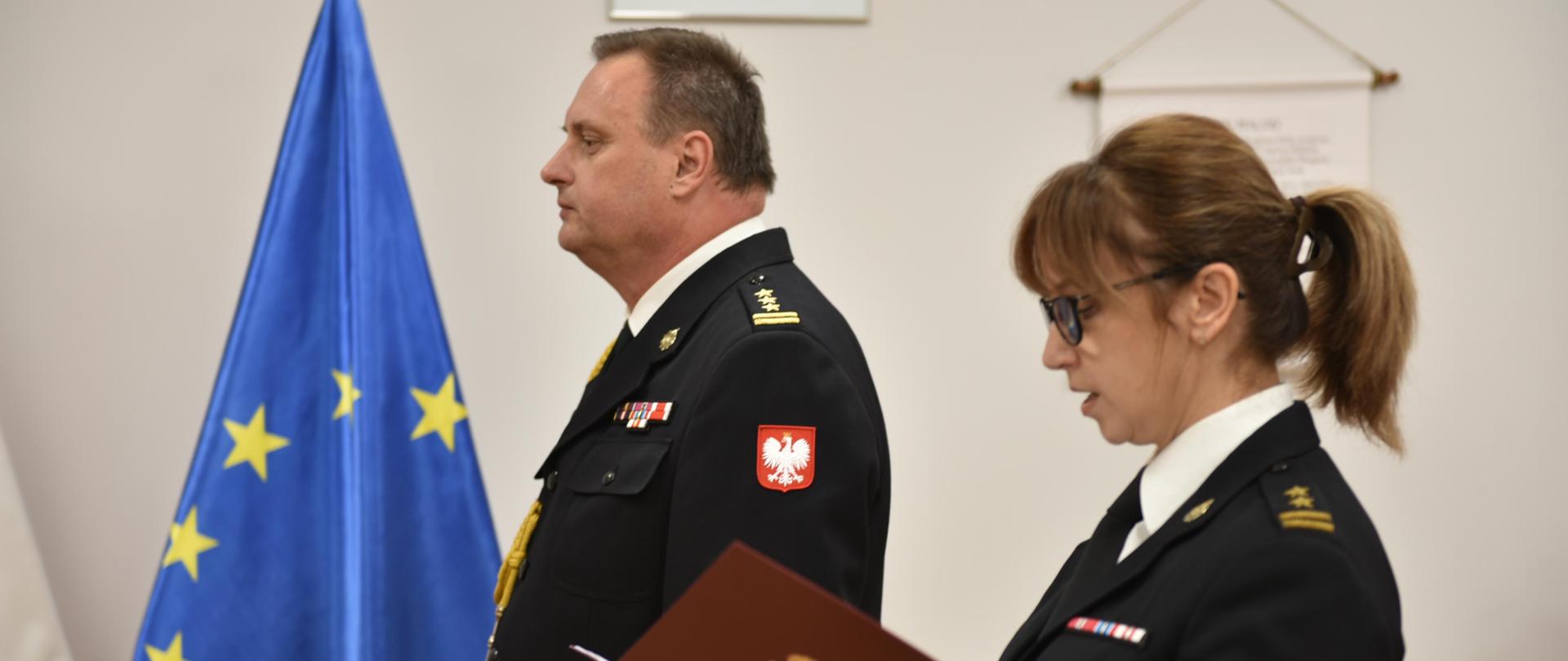 dwóje strażaków stojących obok siebie skierowanych w lewa stronę, funkcjonariuszka po prawej trzyma otwartą bordową teczkę na dokumenty, za nimi flagi polski i unii europejskiej