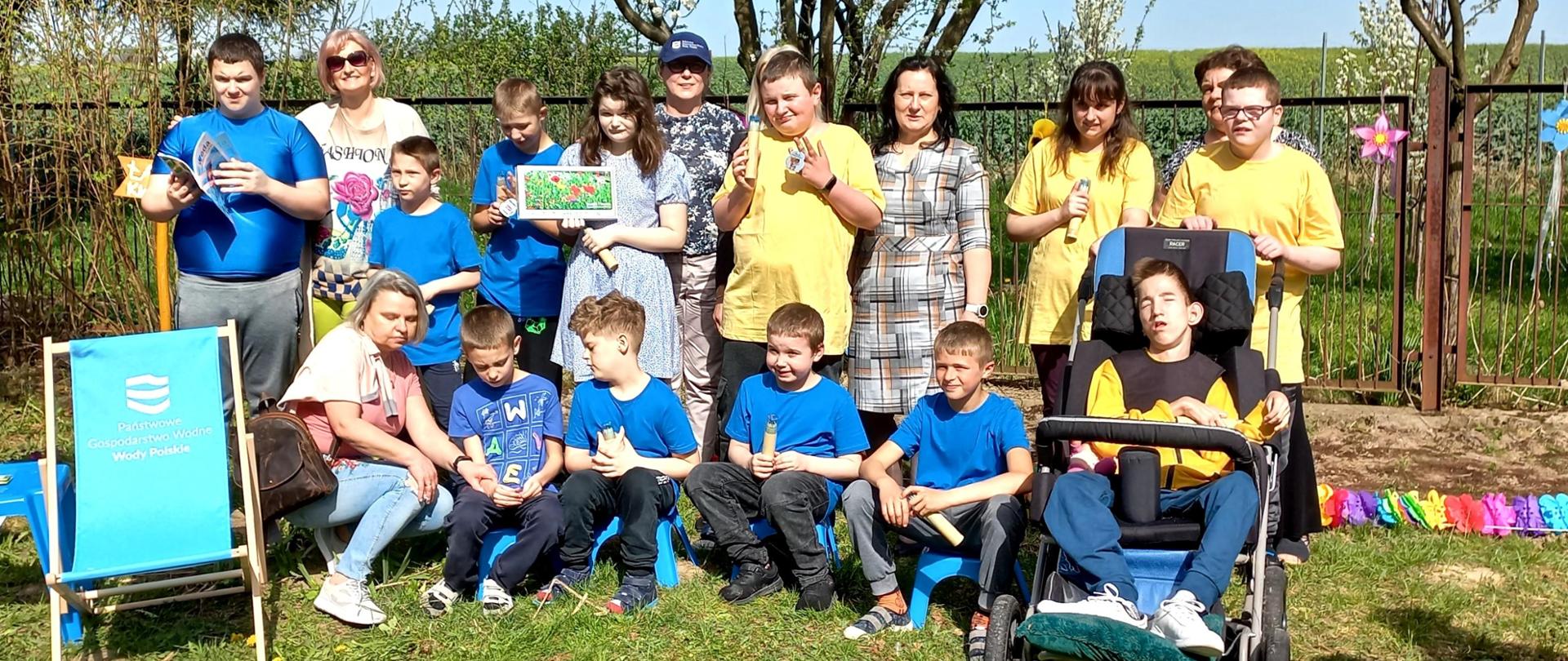Grupa dzieci wraz z nauczycielami i pracownikiem PGW Wody Polskie stoi na trawie w ogródku szkolnym. W tle - kwitnące drzewa.