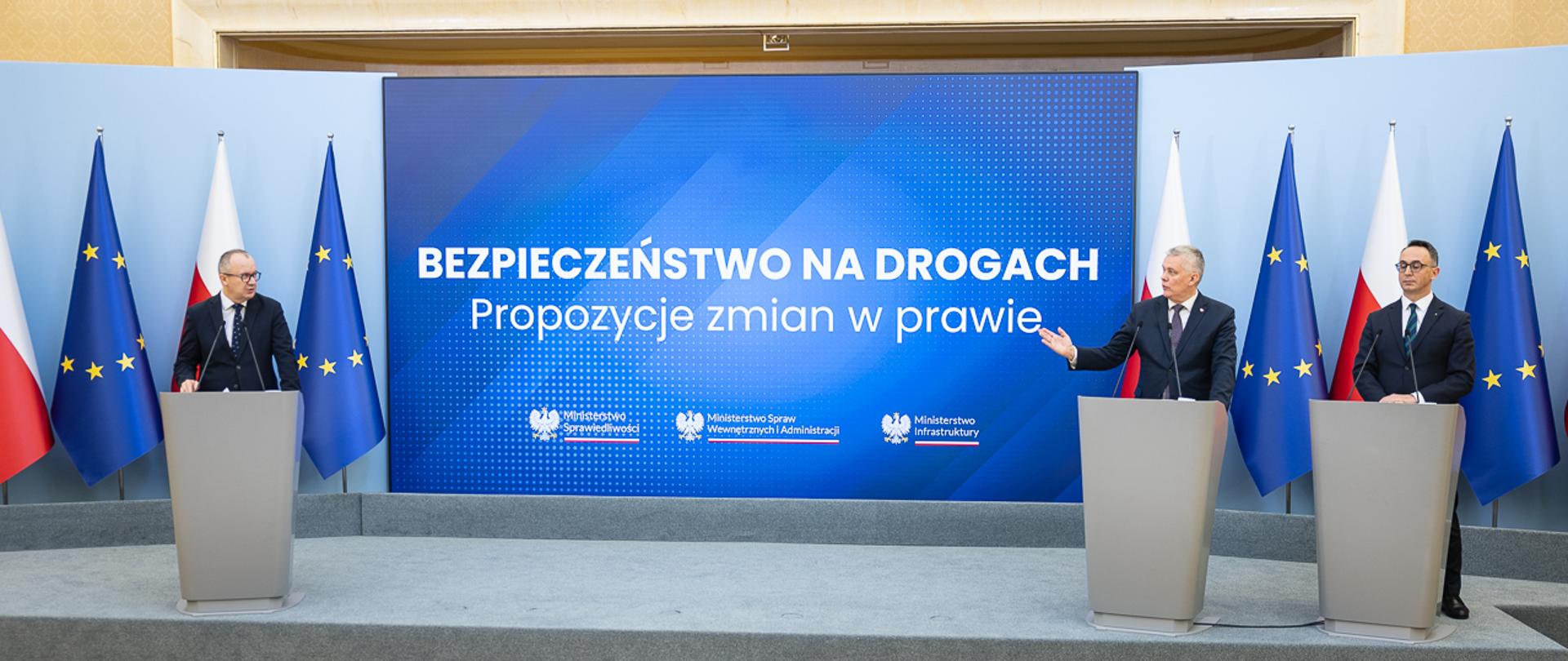 Sala konferencyjna, trzech mężczyzn za mównicami na tle flag Polski i UE na konferencji "Bezpieczeństwo na drogach".