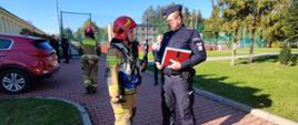 Widok z przodu. Kierujący działaniem ratowniczym strażak w trakcie wspólnych ustaleń z kierującym akcją policjantem. Funkcjonariusze stoją na zewnątrz budynku szkoły, w tle boisko szkolne. 