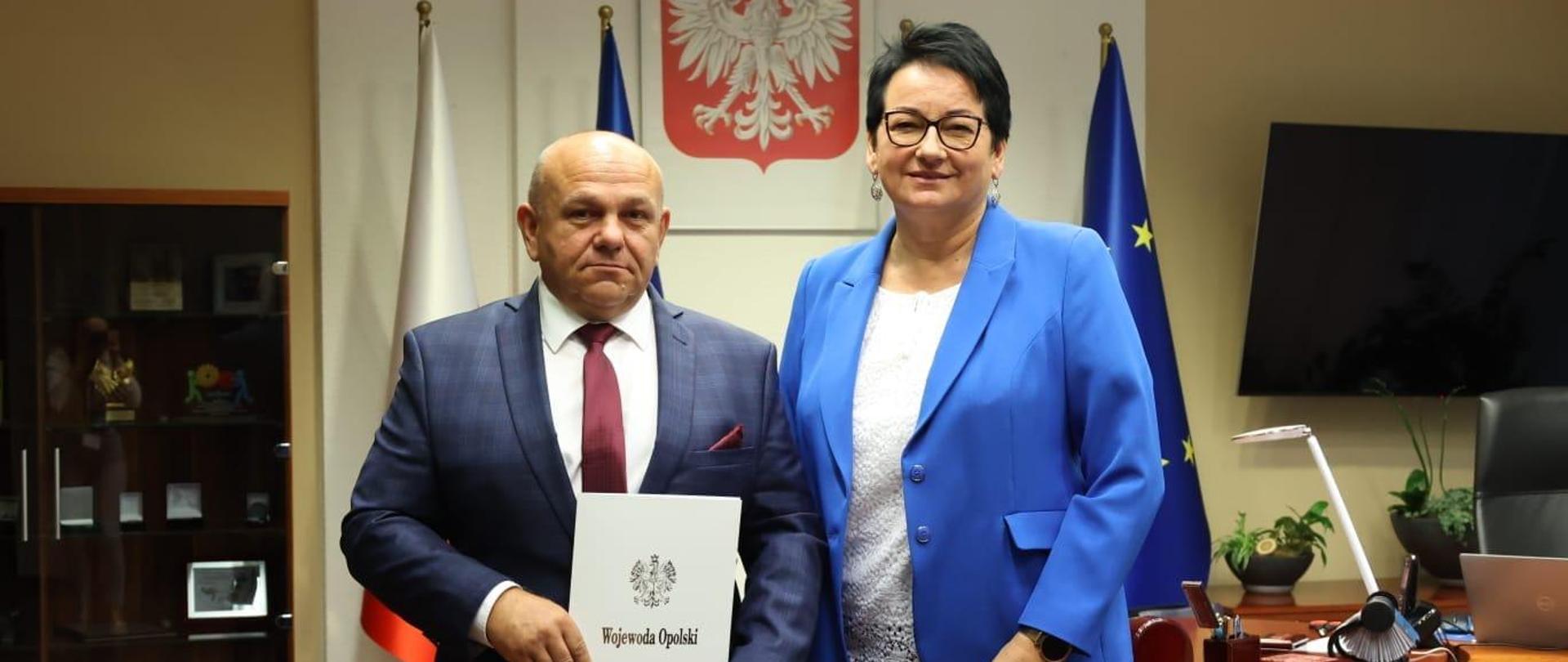 Nowy pełnomocnik oraz Wojewoda pozują do wspólnego zdjęcia. Znajdują się w gabinecie Wojewody. W tle widać biuro oraz przeszkolną szafkę z różnymi nagrodami i podziękowaniami. Pełnomocnik ma w rękach akt powołania. 