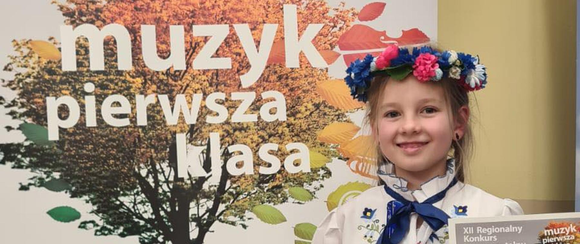 Dziewczynka stoi obok plakatu reklamującego konkurs. W lewej dłoni trzyma dyplom a w prawej nagrodę - statuetkę. 