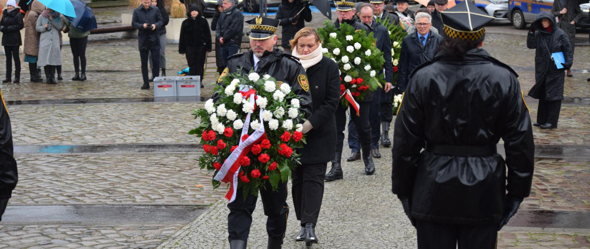 kilka osób idzie w stronę pomnika trzymając wieńce biało czerwonego koloru 