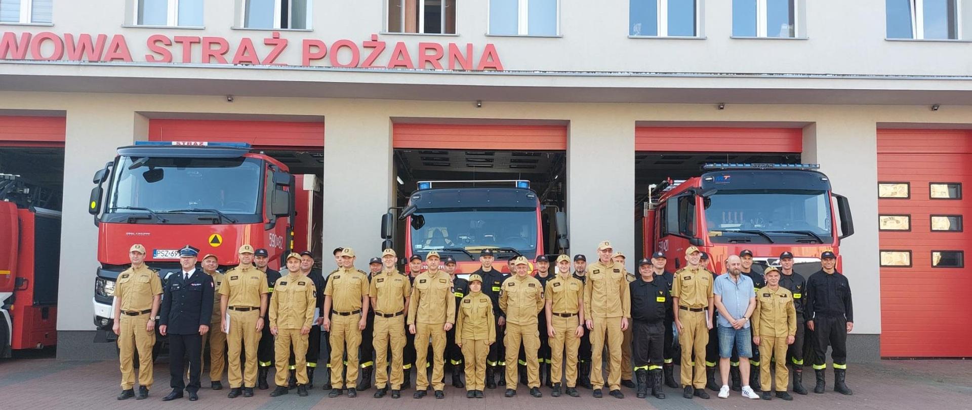 Strażacy na zdjęciu grupowym na tle samochodów i komendy