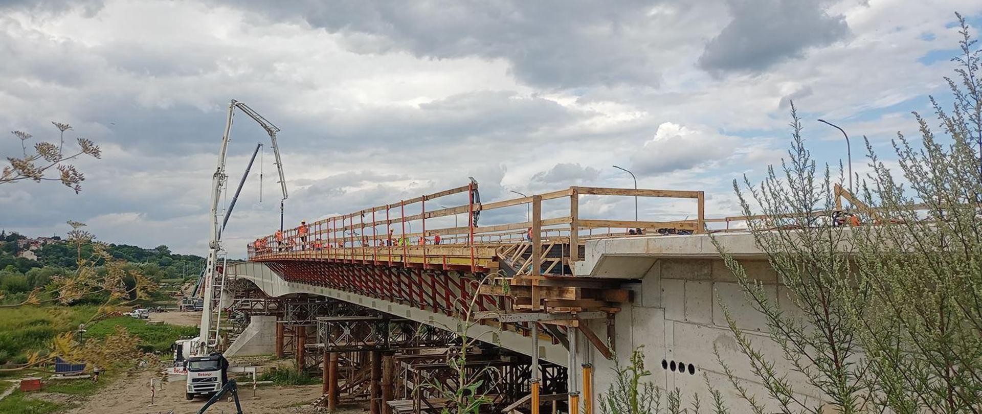 Budowa mostu w Sandomierzu - betonowanie, na zdjęciu konstrukcja na podporach tymczasowych z deskowaniem wykonanym wzdłuż obiektu. Przy moście betoniarka z wysięgnikiem sięgającym do mostu. Na moście pracownicy 