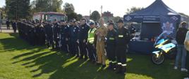 Stadion w Miejskiej Górce. Na tle wozu strażackiego oraz namiotu promocyjnego Komendy Wojewódzkiej Policji w Poznaniu stoją goście, którzy uczestniczą w uroczystej inauguracji wydarzenia. Wśród nich są strażacy i policjanci. Widoczna jest też klasa mundurowa. Obok namiotu zaparkowany jest motocykl policyjny. 