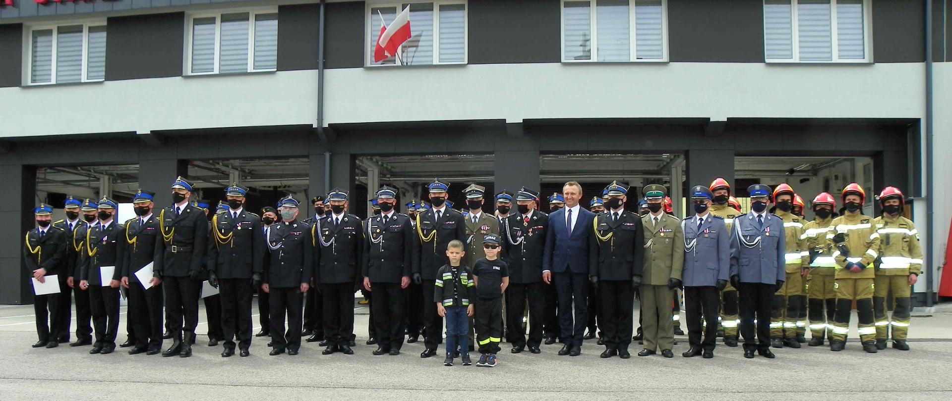 zdjęcie zrobione w dzień. Na zdjęciu widać grupę strażaków, policjantów, wojskowych i zaproszonych gości stojących na placu zewnętrznym przed budyniem JRG Sandomierz, którym jest wykonywane wspólne zdjęcie grupowe. Na budynku JRG widać powiewające flagi państwowe i napis straż pożarna, 998 i 112.