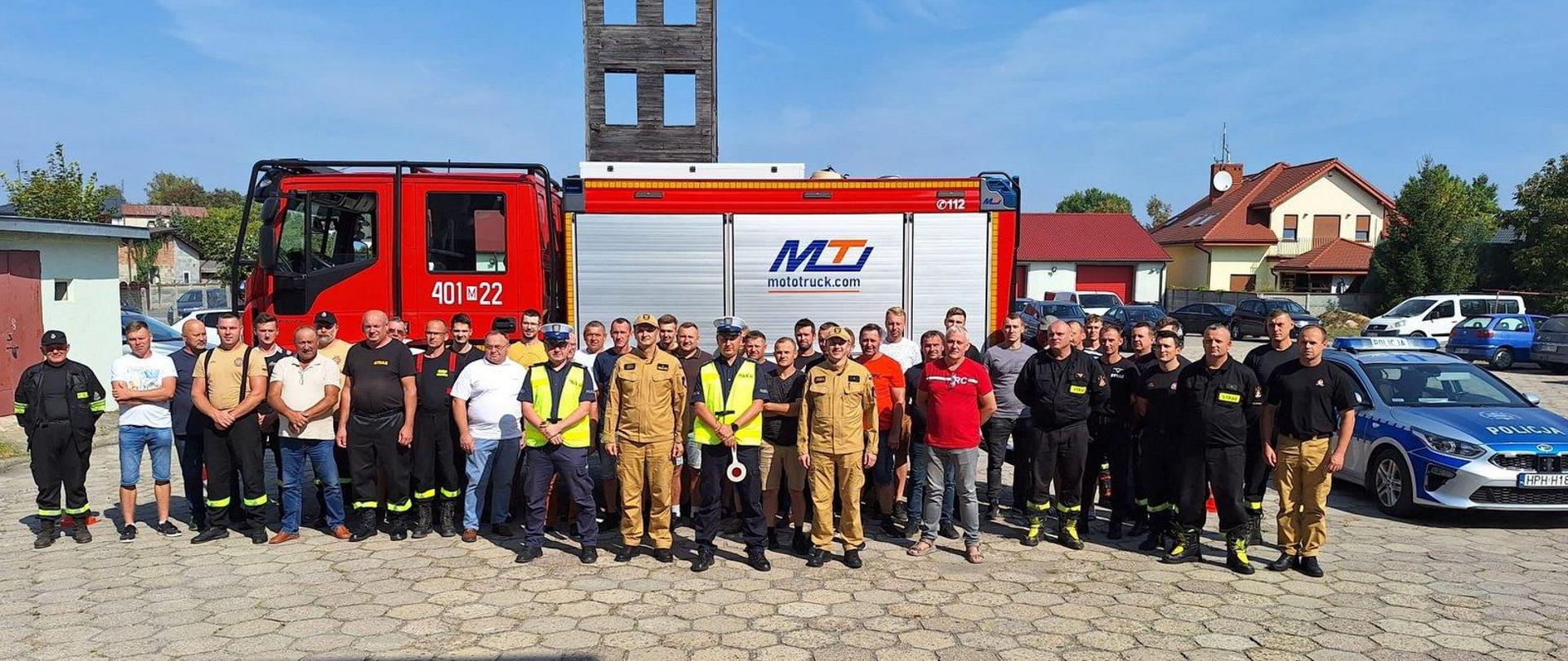 Grupa czterdziestu kierowców przed samochodem pożarniczym. W środku dwóch policjantów i dwóch strażaków. Jeden policjant trzyma "lizak ostrzegawczy". Z prawej strony radiowóz policyjny.