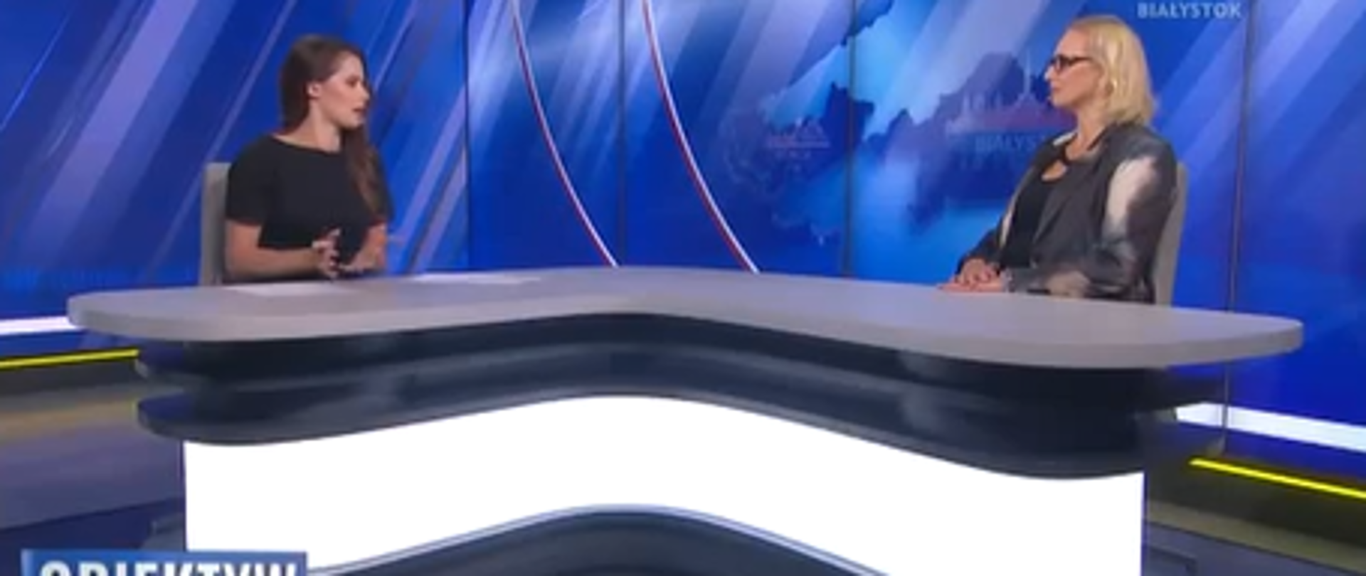 Fotografia dwóch pań siedzących naprzeciw siebie w studio telewizyjnym TVP3 Białystok i rozmawiających na temat zbierania grzybów. Kolorystyka niebiesko-szaro-biała.