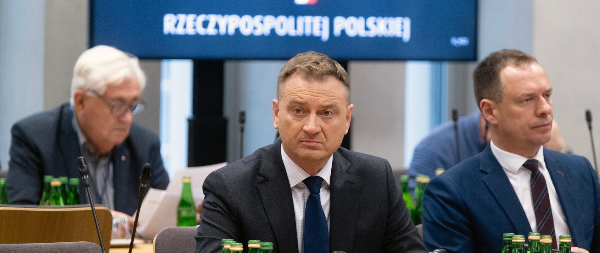 Minister Sportu i Turystyki Sławomir Nitras siedzi za stołem podczas posiedzenia Komisji Kultury Fizycznej, Sportu i Turystyki. Przed nim na stole stoją butelki z wodą. Obok niego siedzi Sekretarz Stanu Piotr Borys. Za nimi drugi stół. W tle napis na granatowym tle "Sejm Rzeczypospolitej Polskiej".