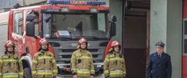 Strażacy ubrani w ubrania specjalne oraz komendant powiatowy bryg. Andrzej Wójtowicz w płaszczu wyjściowym zimowym stoją na tle samochodów ratowniczo-gaśniczych